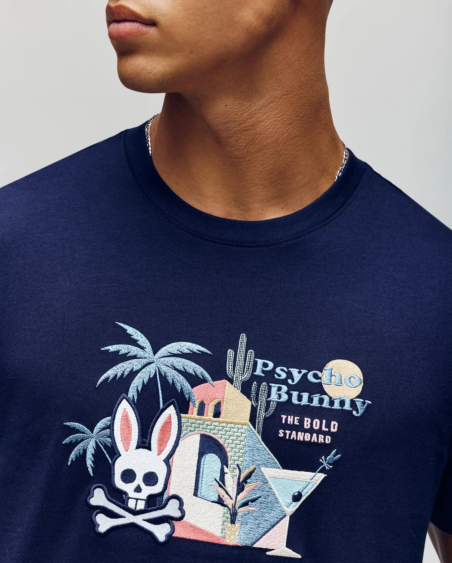 A person wears the Psycho Bunny MENS SHANE GRAPHIC TEE (MT0200175), a navy summer tee in soft Pima cotton with colorful embroidered tropical graphics—palm tree, cactus, building, cocktail, and bunny skull logo. Text: "Psycho Bunny THE BOLD STANDARD.