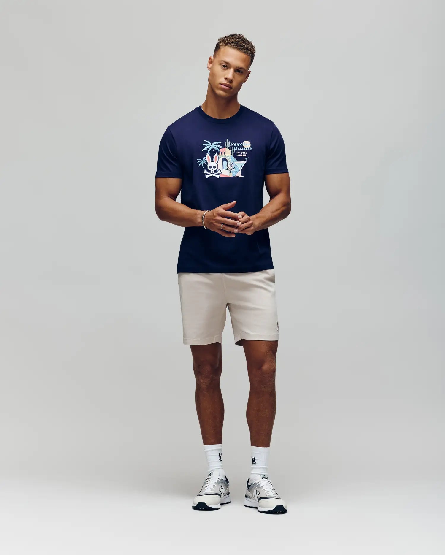 A man stands against a plain background wearing the Psycho Bunny MENS SHANE GRAPHIC TEE - MT0200175, paired with light beige shorts, white socks, and white sneakers. He stands with hands clasped, looking forward in a relaxed pose.