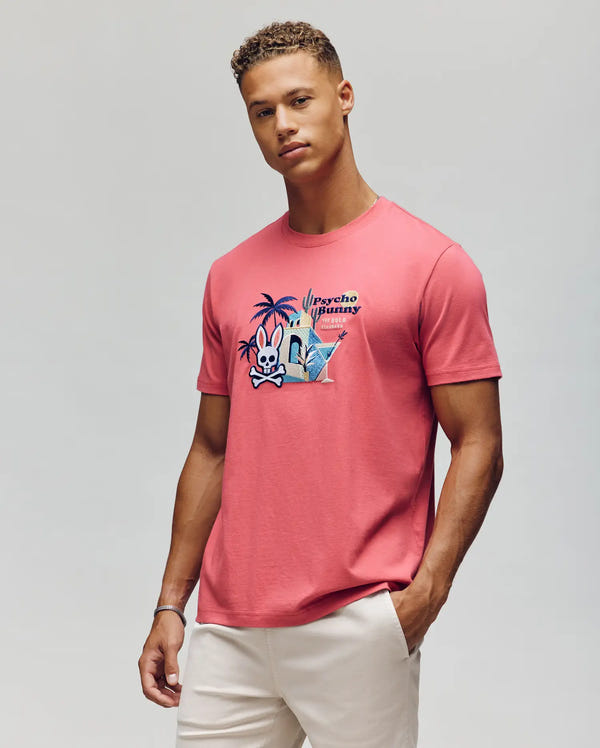 A man wearing the Psycho Bunny MENS SHANE GRAPHIC TEE - MT0200175, a coral pink Pima cotton t-shirt with cartoon palm trees, Martini glasses, and a bunny on the front, paired with light beige pants, stands against a gray background.