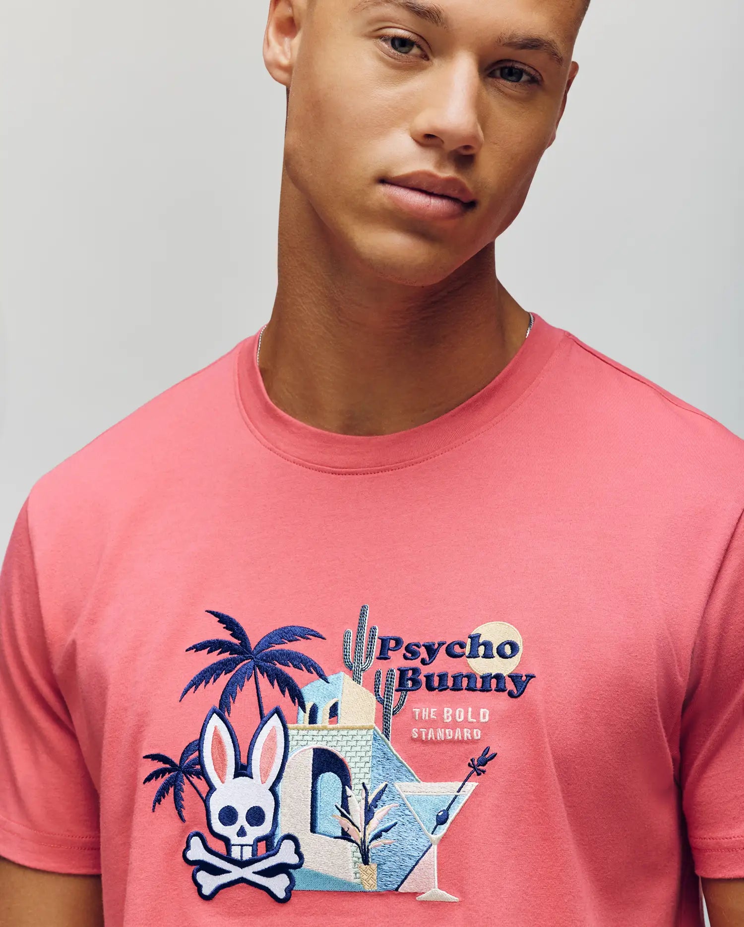 A young man wears the MENS SHANE GRAPHIC TEE (MT0200175) by Psycho Bunny, a pink Pima cotton summer tee with a desert scene, palm trees, martini glasses, and the signature white bunny skull-and-crossbones logo.