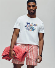 A man wears the Psycho Bunny MENS SHANE GRAPHIC TEE (MT0200175), a white Pima cotton shirt, paired with red-and-white striped shorts. Holding a folded red towel, he poses against a light background with a relaxed, summer-ready vibe.