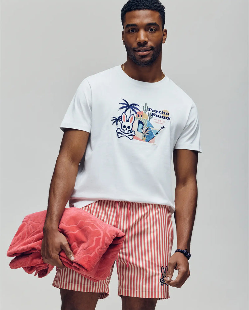A man wears the Psycho Bunny MENS SHANE GRAPHIC TEE (MT0200175), a white Pima cotton shirt, paired with red-and-white striped shorts. Holding a folded red towel, he poses against a light background with a relaxed, summer-ready vibe.