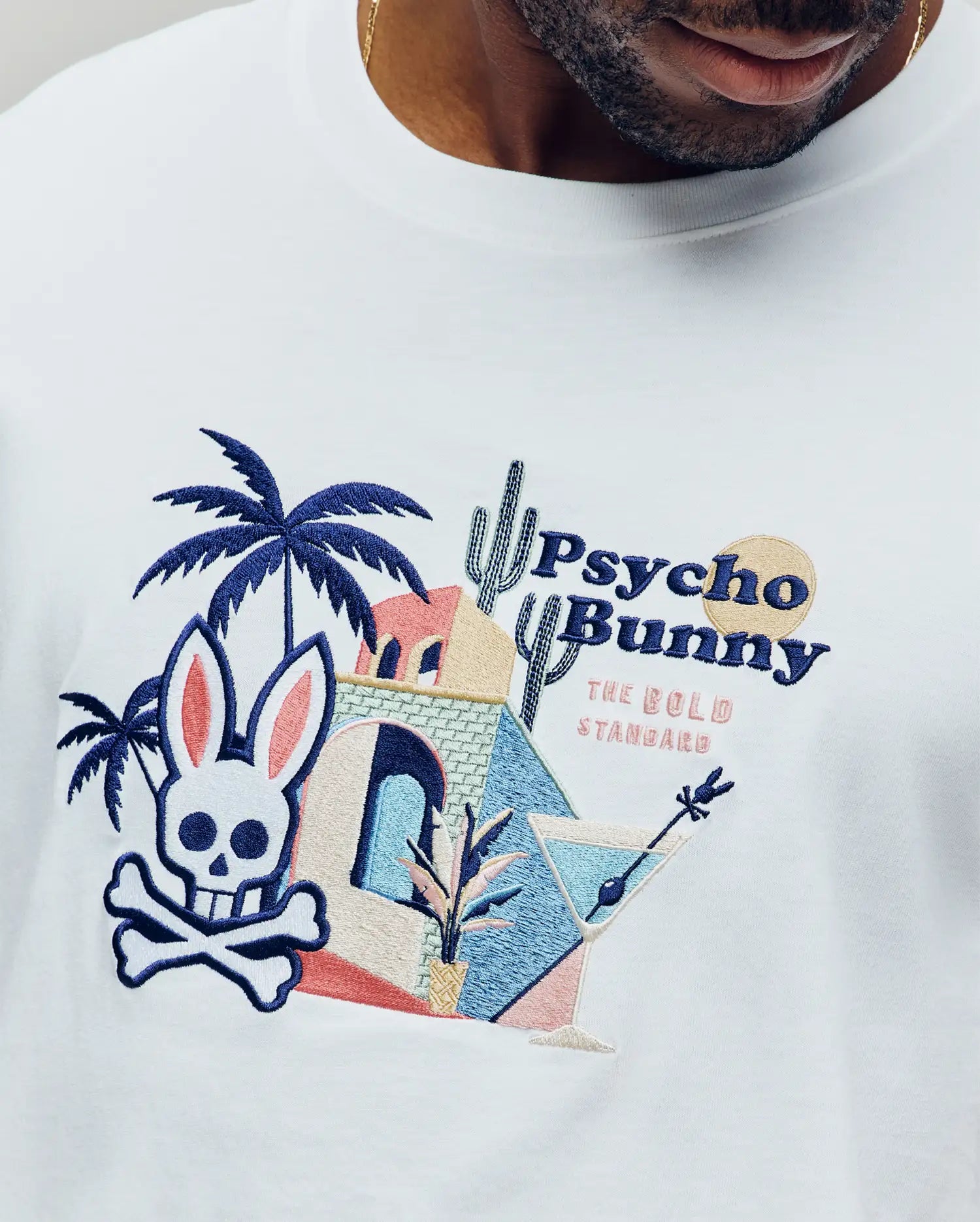 A man wears the Psycho Bunny MENS SHANE GRAPHIC TEE (MT0200175), a white Pima cotton shirt with a vivid embroidered skull bunny, palm trees, cacti, and "Psycho Bunny The Gold Standard"—perfect for summer.