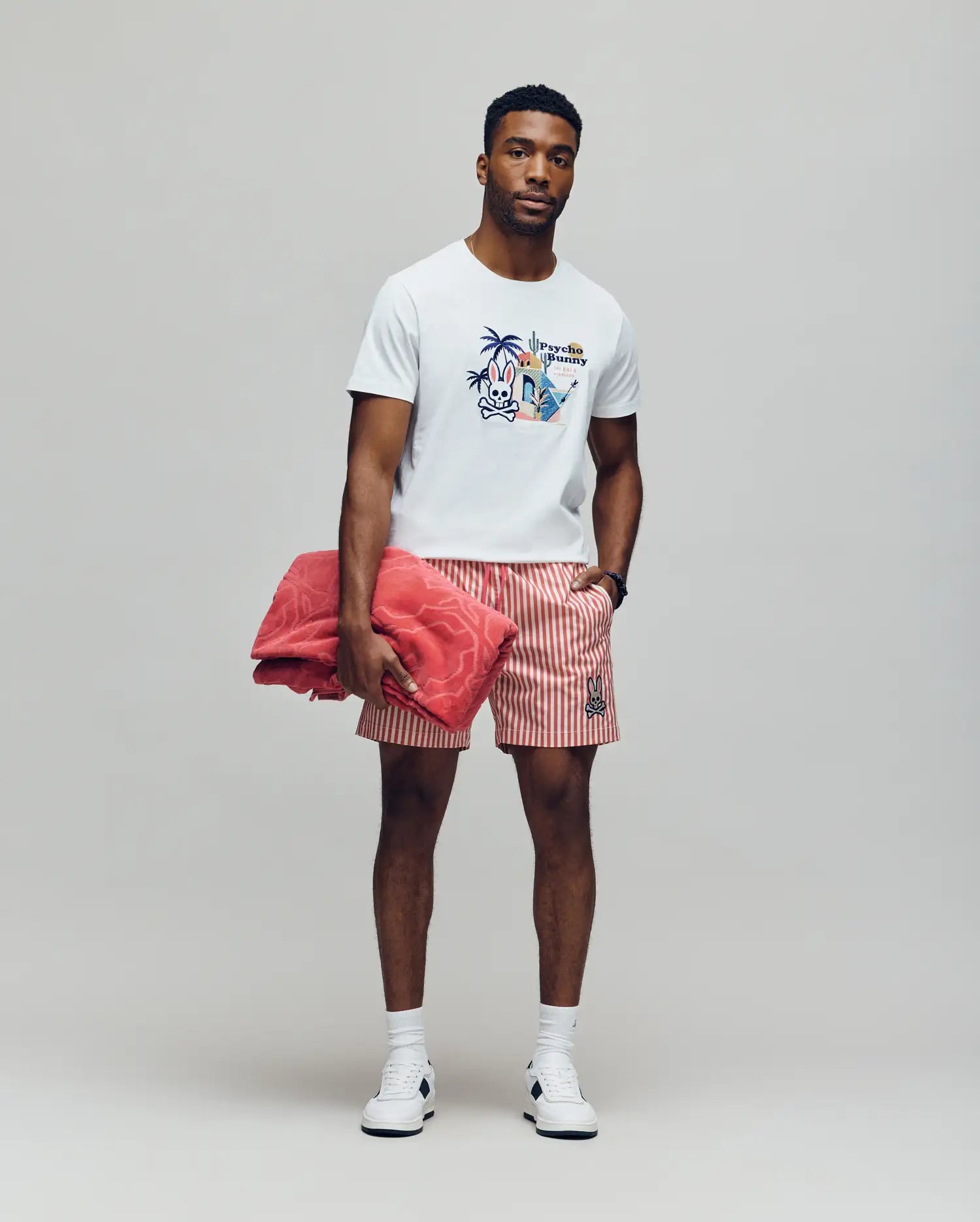 A man stands against a plain background wearing the Psycho Bunny MENS SHANE GRAPHIC TEE - MT0200175, red and white striped shorts, white sneakers, and a watch. He holds a folded pink towel under one arm and looks calmly at the camera.