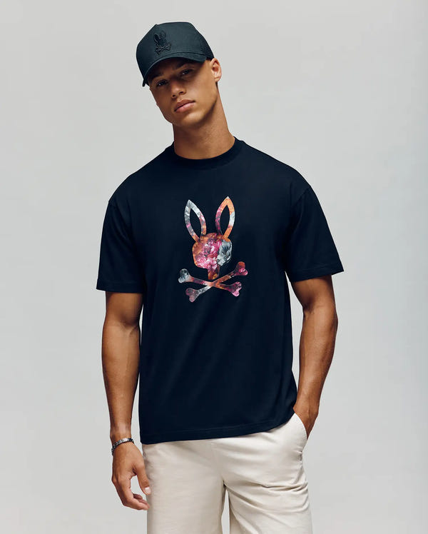 A man in a black cap, light pants, and the Psycho Bunny MENS NOVAN GRAPHIC TEE (MT0200177)—a black Pima cotton shirt with a colorful bunny and crossbones—stands with one hand in his pocket against a plain background.