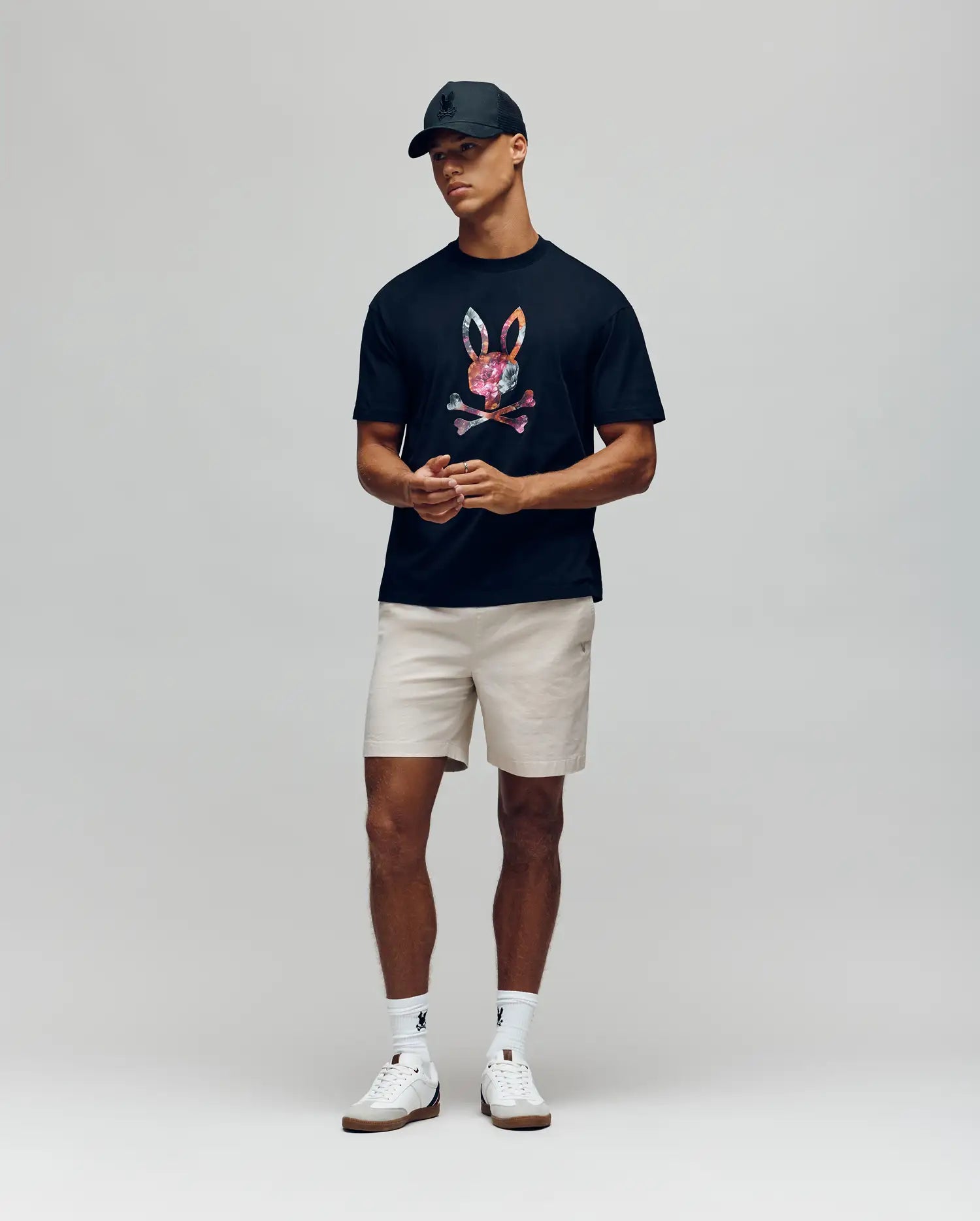 A man in a black cap, beige shorts, white socks and sneakers wears the Psycho Bunny MENS NOVAN GRAPHIC TEE (MT0200177), featuring a colorful bunny print, standing against a plain light gray background.