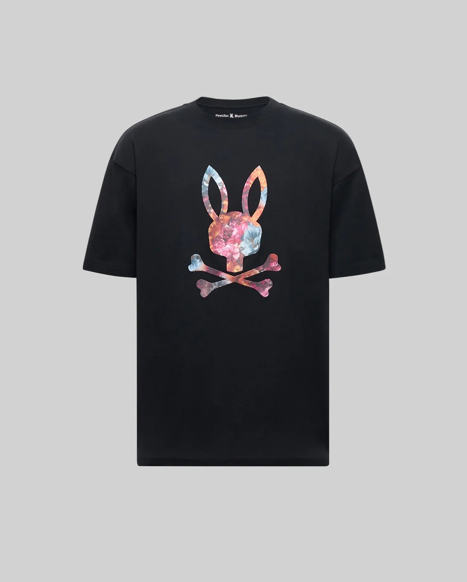 The Psycho Bunny MENS NOVAN GRAPHIC TEE - MT0200177 is a black Pima cotton shirt featuring a vibrant tie-dye style Printed Bunny with upright ears above crossed bones on the chest, set against a gray background.