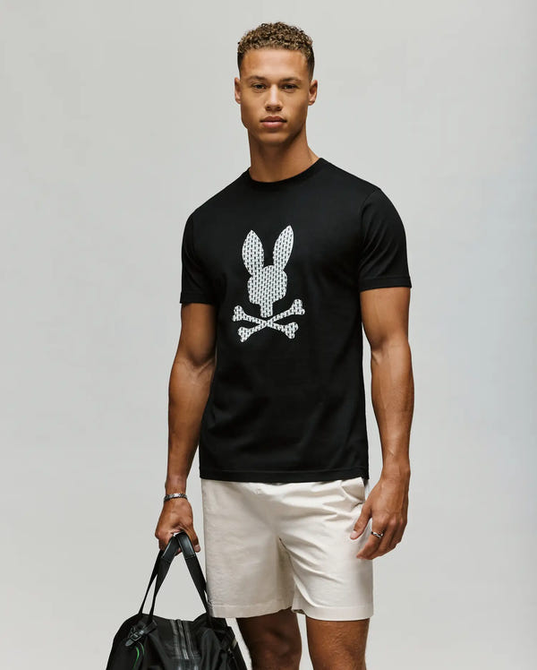 A man stands against a plain background, wearing the Psycho Bunny MENS TALON GRAPHIC TEE - MT0200179 in black with a white bunny and crossbones graphic, paired with white shorts and holding a black duffel bag in his left hand.