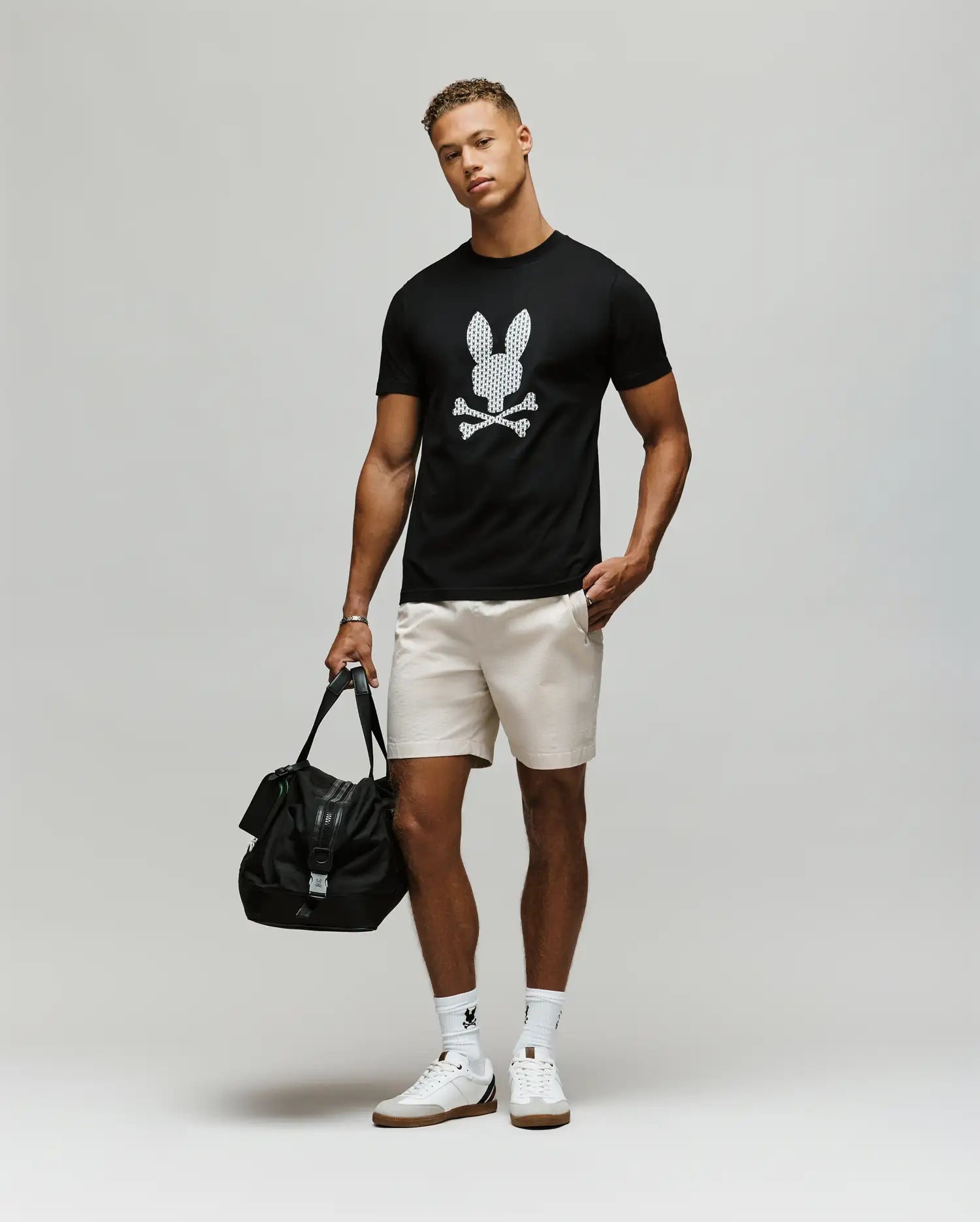 A man wears the Psycho Bunny MENS TALON GRAPHIC TEE (MT0200179), a regular fit black 100% cotton tee with a graphic bunny skull, paired with light shorts, white socks, white sneakers, and holds a black bag in his left hand.