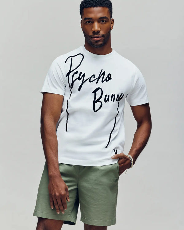 A man wears a white Psycho Bunny MENS SOREN GRAPHIC TEE (MT0200180), made of 100% cotton with a flocked "Psycho Bunny" wordmark, and light green shorts, standing against a plain light gray background.