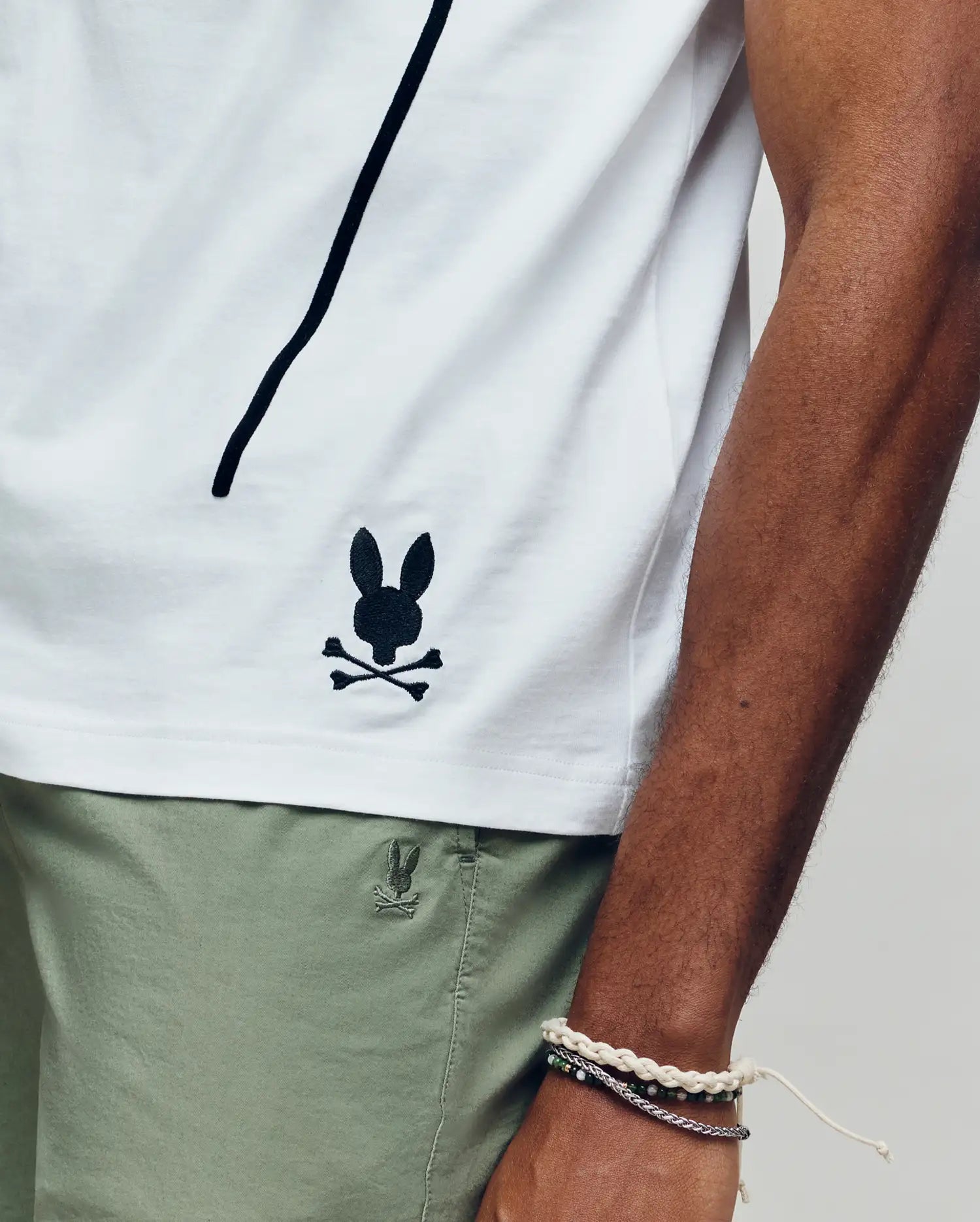 Wearing the Psycho Bunny MENS SOREN GRAPHIC TEE (MT0200180), a 100% cotton white tee with a black bunny and crossbones logo near the hem, and light green shorts, they rest their bracelet-adorned arm along the shirt’s side seam.
