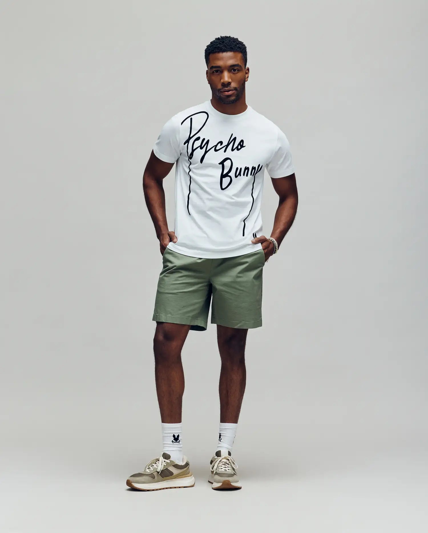 A man stands against a plain background wearing the Psycho Bunny MENS SOREN GRAPHIC TEE (MT0200180) in white, paired with light green shorts, white socks, and beige sneakers. His hands are in his pockets as he looks straight ahead.