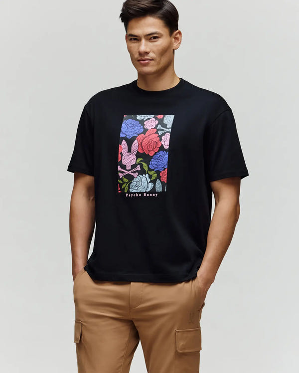 A man wears the Psycho Bunny MENS ROWEN OVERSIZED TEE (MT0200194), a relaxed fit black Pima cotton shirt with a colorful floral graphic and "Psycho Bunny" text, paired with tan cargo pants, standing against a light gray background.