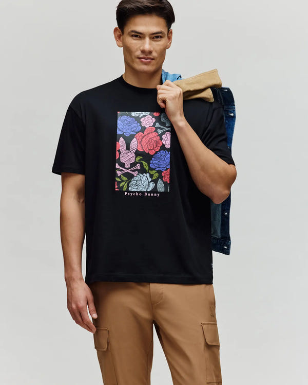 A man wears the Psycho Bunny MENS ROWEN OVERSIZED TEE - MT0200194, a relaxed-fit black Pima cotton shirt with a bold floral graphic and logo. He pairs it with tan cargo pants, holding a denim jacket and tan garment over his shoulder against a plain background.