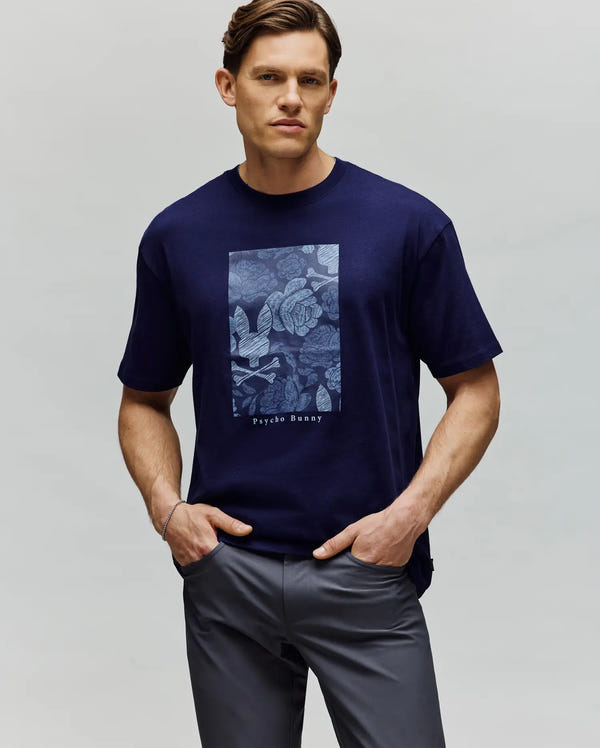 A man stands with hands in pockets against a plain light gray background wearing the Psycho Bunny MENS ROWEN OVERSIZED TEE (MT0200194), a navy blue relaxed-fit shirt featuring a rectangular blue floral design and "Psycho Bunny" text.