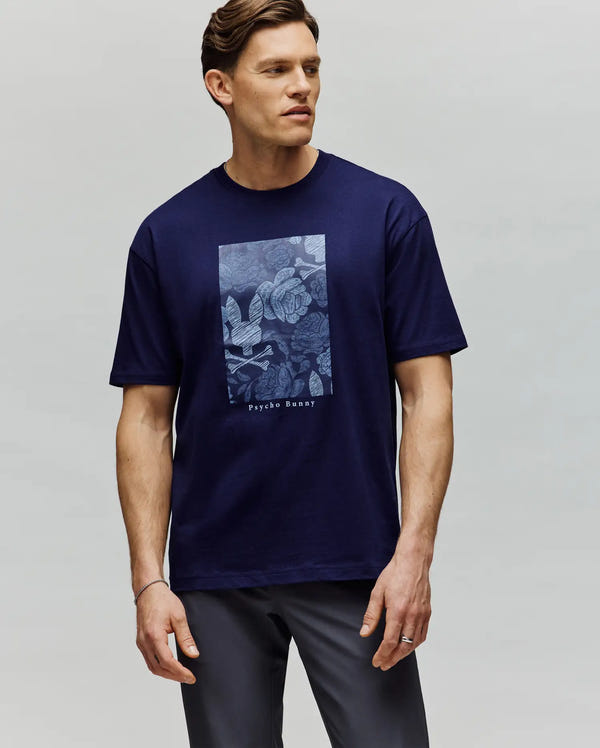 A man with short brown hair stands against a light gray background, wearing the Psycho Bunny MENS ROWEN OVERSIZED TEE (MT0200194)—a navy blue Pima cotton tee with a light blue floral graphic and "Psychic Beauty" text. He looks slightly to the side.