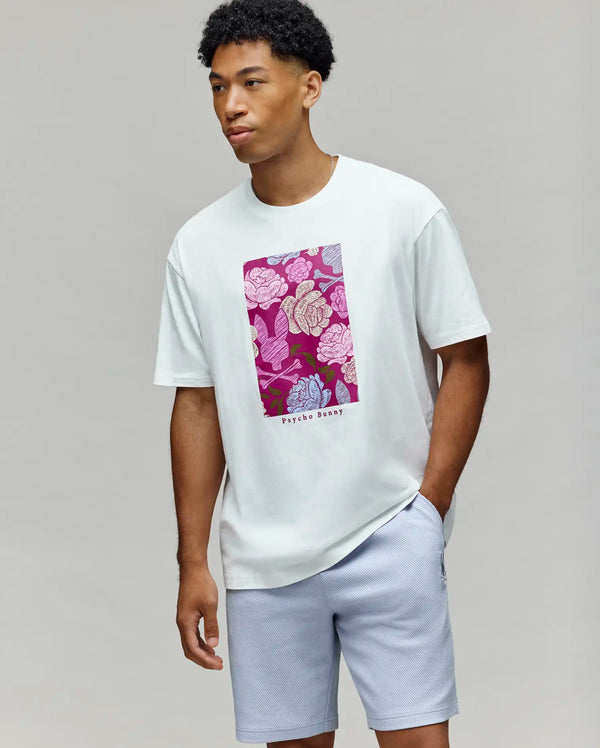 A man stands against a light gray background, wearing the Psycho Bunny MENS ROWEN OVERSIZED TEE (MT0200194) with a vibrant floral graphic and logo, paired perfectly with light gray shorts.