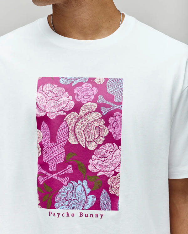 A person wears the Psycho Bunny MENS ROWEN OVERSIZED TEE (MT0200194), a relaxed fit t-shirt featuring a rectangular floral graphic in pink, blue, and white with bunny logos and "Psycho Bunny" printed below.