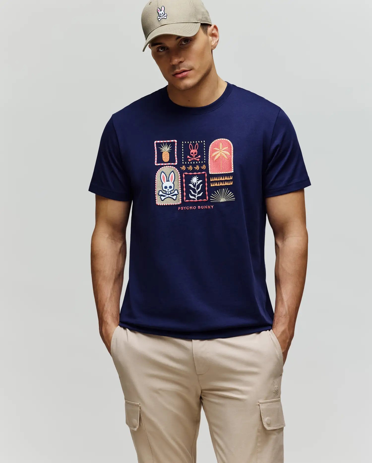 A man wears a beige cap and Psycho Bunny MENS SUNDEW GRAPHIC TEE (MT0200195), a navy Pima cotton shirt with colorful Beach Bunny and palm tree patches, paired with light beige cargo pants, standing against a plain background.