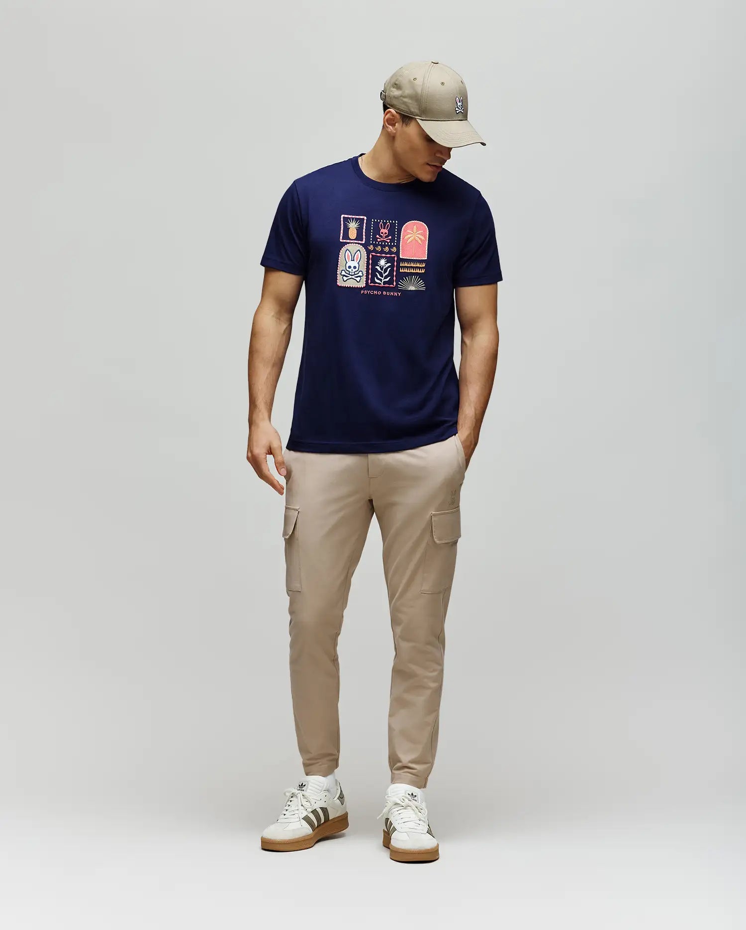 A man wears the Psycho Bunny MENS SUNDEW GRAPHIC TEE (MT0200195) in navy, paired with beige cargo pants, white sneakers, and a beige cap. He stands against a light gray background, looking down with his hands in his pockets.