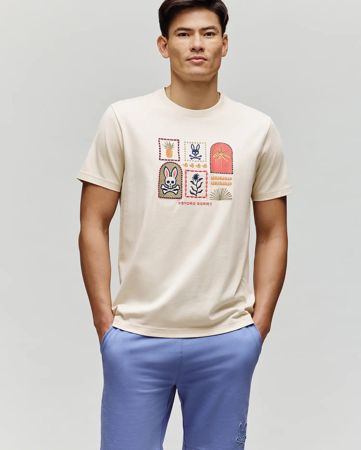 A man wears the Psycho Bunny MENS SUNDEW GRAPHIC TEE (MT0200195), featuring colorful graphics, paired with blue shorts and posing against a light background for a breezy, ocean-inspired look.