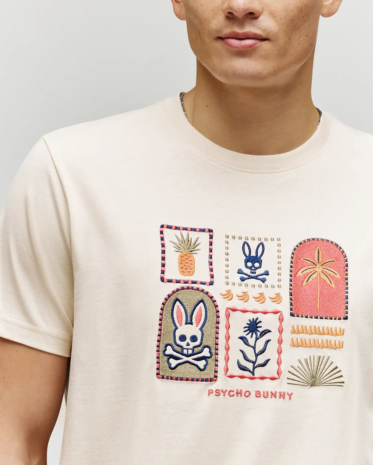A person wears the Psycho Bunny MENS SUNDEW GRAPHIC TEE (MT0200195), a cream Pima cotton shirt featuring colorful embroidered patches—a bunny with crossbones, pineapple, flower, palm—and "Psycho Bunny" text for an ocean-view vibe.