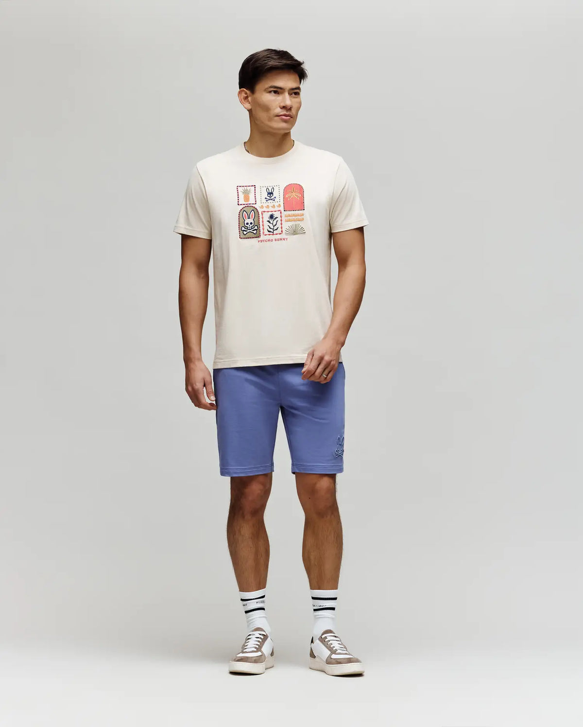 A man stands against a plain background wearing the Psycho Bunny MENS SUNDEW GRAPHIC TEE - MT0200195, light blue shorts, striped white socks, and beige sneakers for an effortless ocean view style.