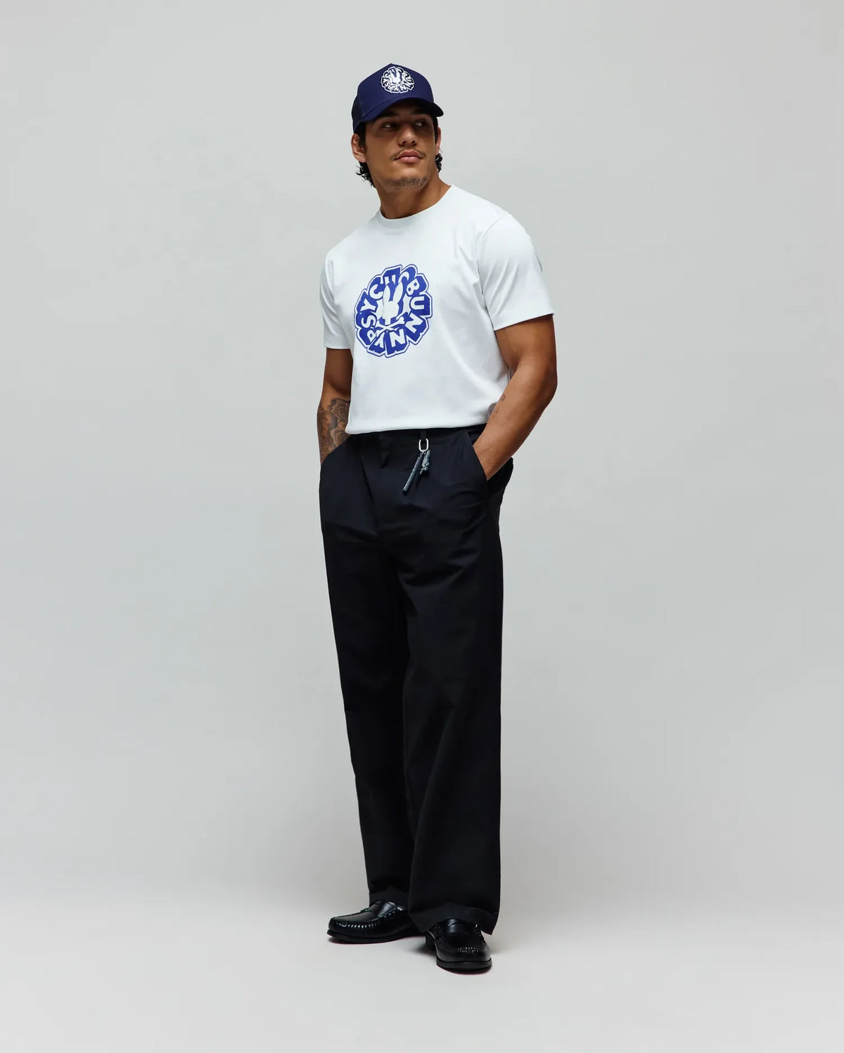 Mens Toby Graphic Tee, a white jersey t-shirt with a large blue Psycho Bunny circular graphic on the chest, paired with relaxed black pants and a navy cap featuring a white embroidered bunny skull.