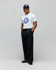 Mens Toby Graphic Tee, a white jersey t-shirt with a large blue Psycho Bunny circular graphic on the chest, paired with relaxed black pants and a navy cap featuring a white embroidered bunny skull.