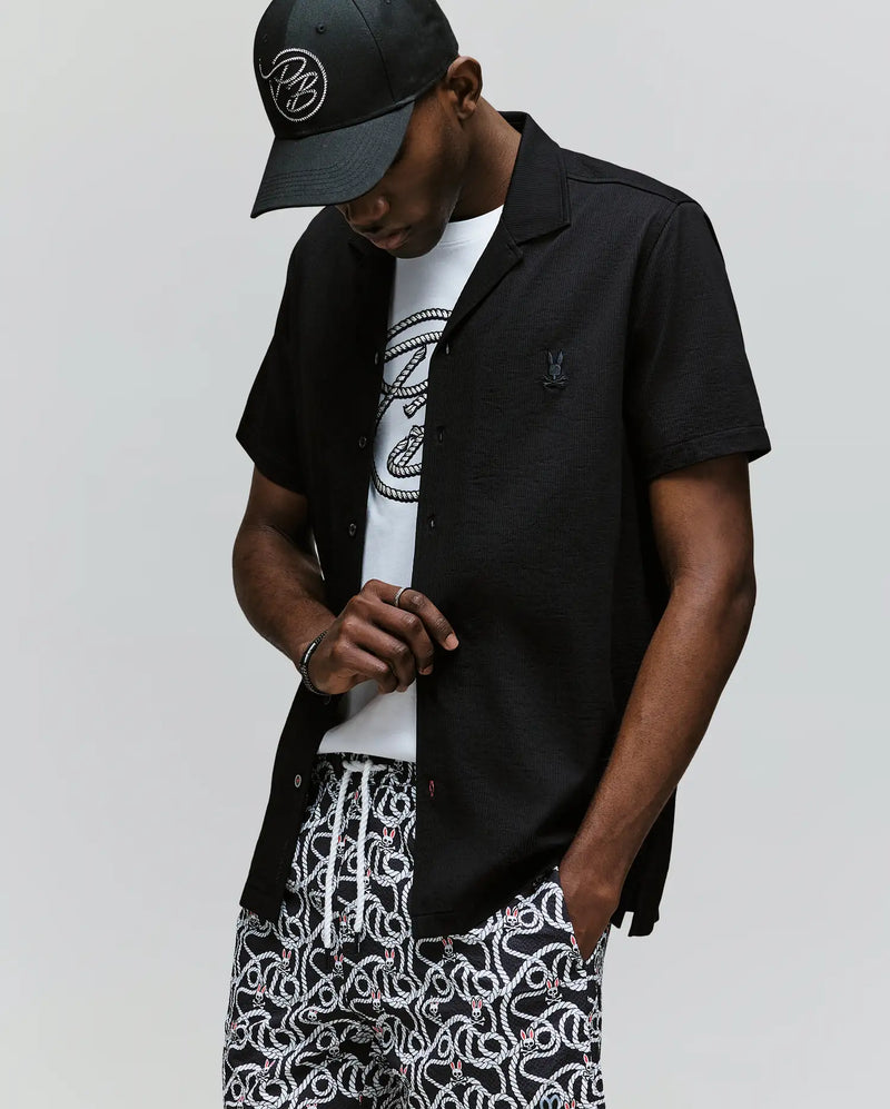A man wears the Psycho Bunny MENS JAYME KNIT SEERSUCKER CABANA COLLAR SHIRT - MT0300002 in black over a white graphic tee, paired with patterned black-and-white shorts and a black cap, standing with his head slightly down and hand touching his shirt.