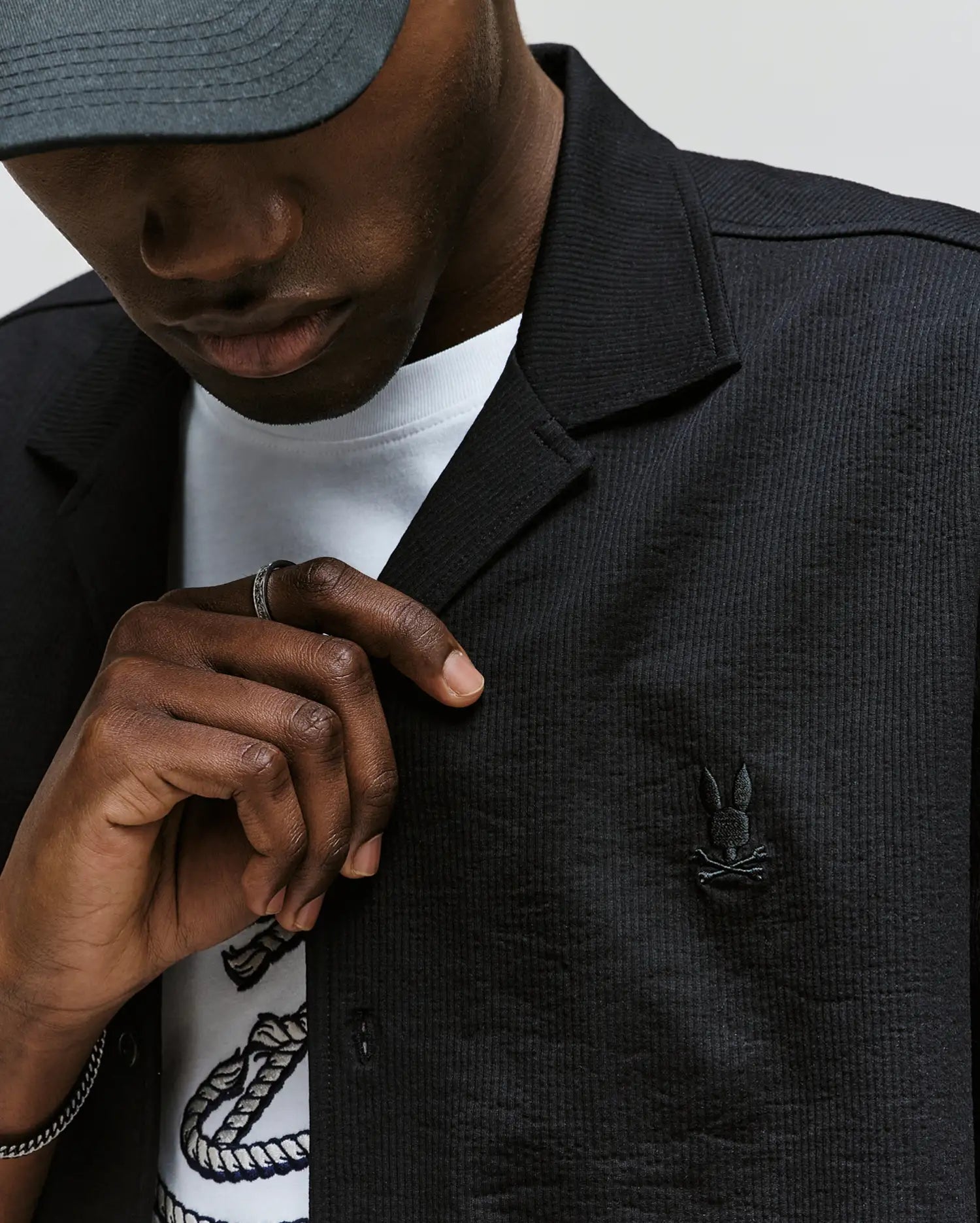 A person wears the Psycho Bunny MENS JAYME KNIT SEERSUCKER CABANA COLLAR SHIRT - MT0300002 with its signature embroidered bunny logo, layered over a white printed tee, paired with a gray cap and ring, gently holding the shirt collar. Their face is partly visible.