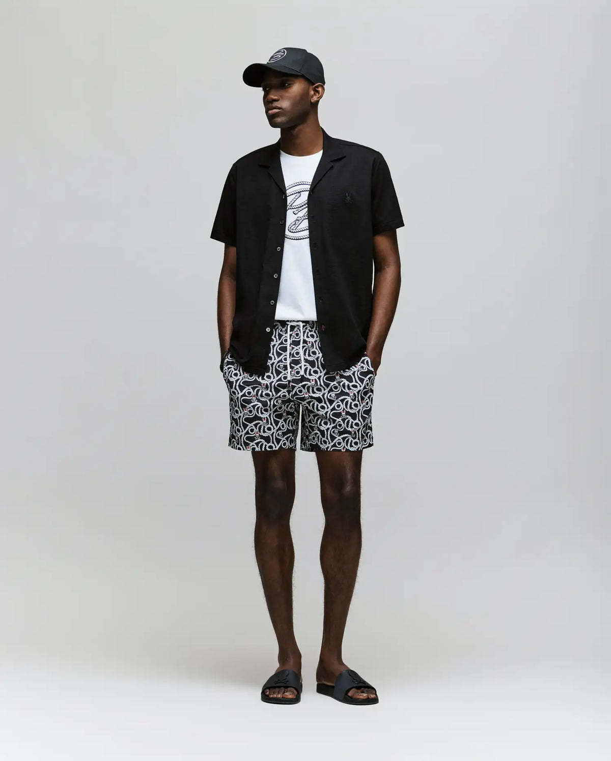 A man stands against a plain background wearing the Psycho Bunny MENS JAYME KNIT SEERSUCKER CABANA COLLAR SHIRT - MT0300002 over a white graphic tee, patterned shorts, and black slide sandals. His hands are in his pockets.