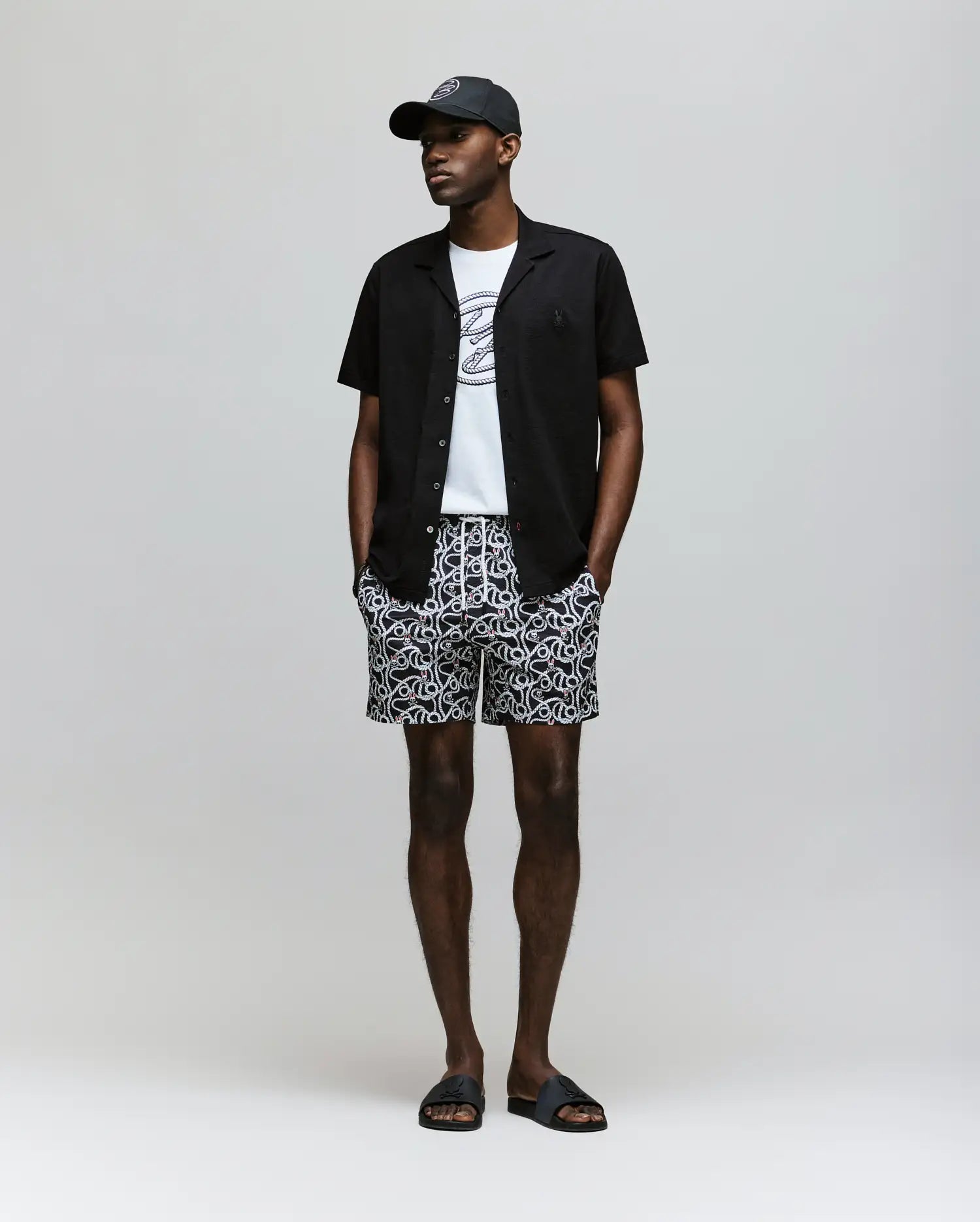 A man stands against a plain background wearing the Psycho Bunny MENS JAYME KNIT SEERSUCKER CABANA COLLAR SHIRT - MT0300002 over a white graphic tee, patterned shorts, and black slide sandals. His hands are in his pockets.