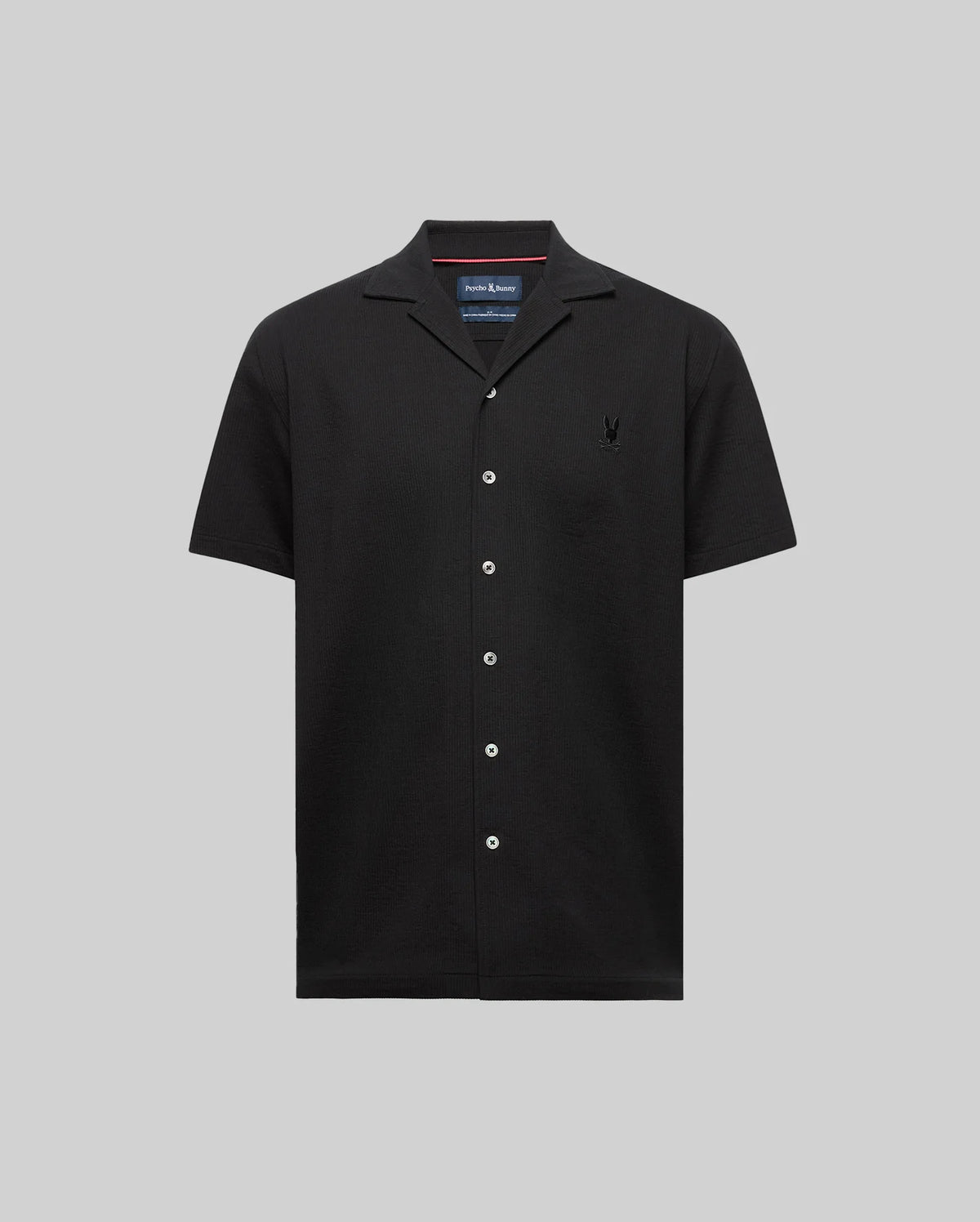 The Psycho Bunny MENS JAYME KNIT SEERSUCKER CABANA COLLAR SHIRT (MT0300002) is a black short-sleeve cotton-blend shirt with a cabana collar, white buttons, and an embroidered logo on the left chest, shown on a light gray background.