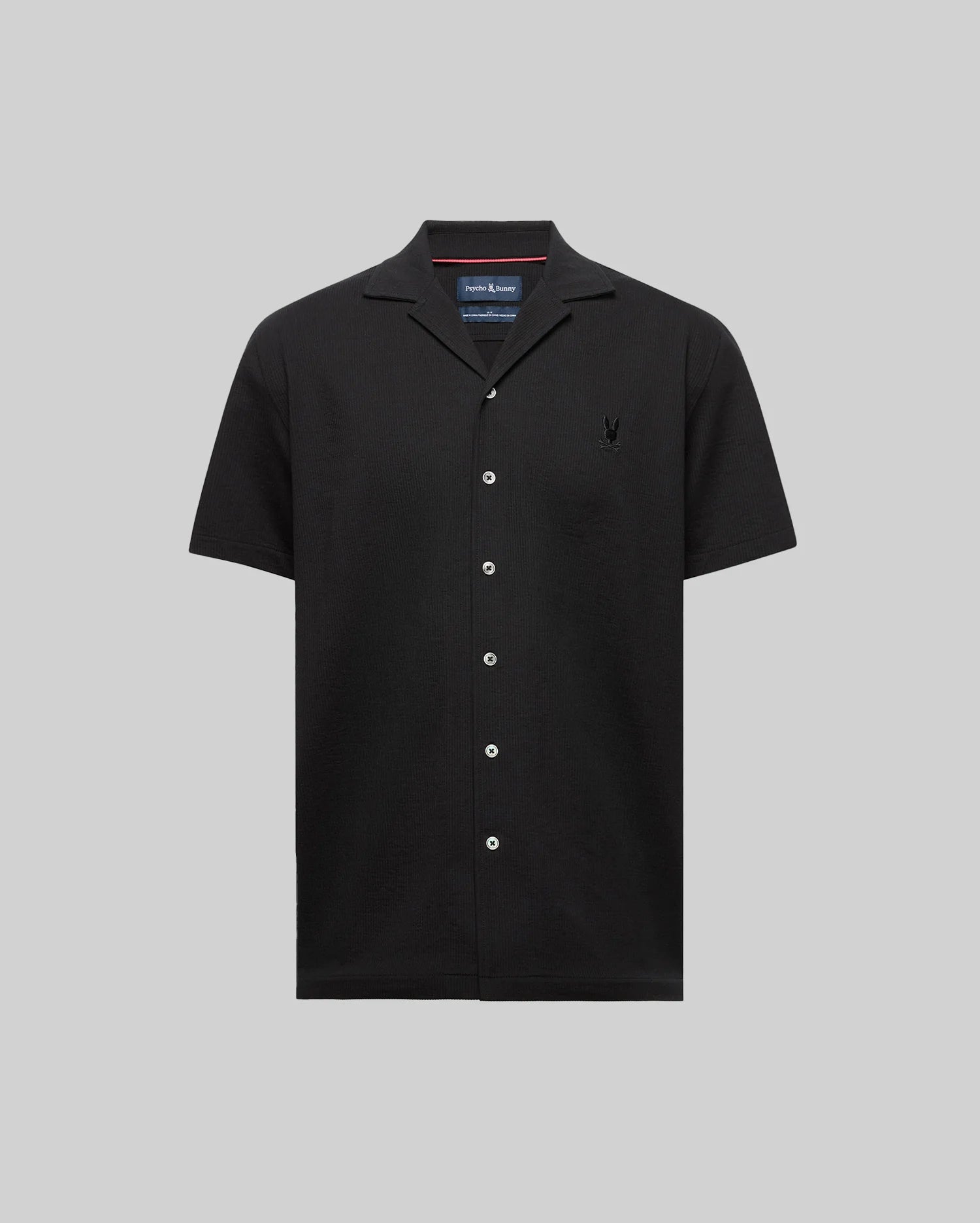 The Psycho Bunny MENS JAYME KNIT SEERSUCKER CABANA COLLAR SHIRT (MT0300002) is a black short-sleeve cotton-blend shirt with a cabana collar, white buttons, and an embroidered logo on the left chest, shown on a light gray background.