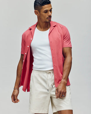 A man stands against a neutral background wearing the PsychoBunny MENS JAYME KNIT SEERSUCKER CABANA COLLAR SHIRT (MT0300002) in pink over a white tank and light beige drawstring shorts, looking down with one hand by his side, the other on his thigh.