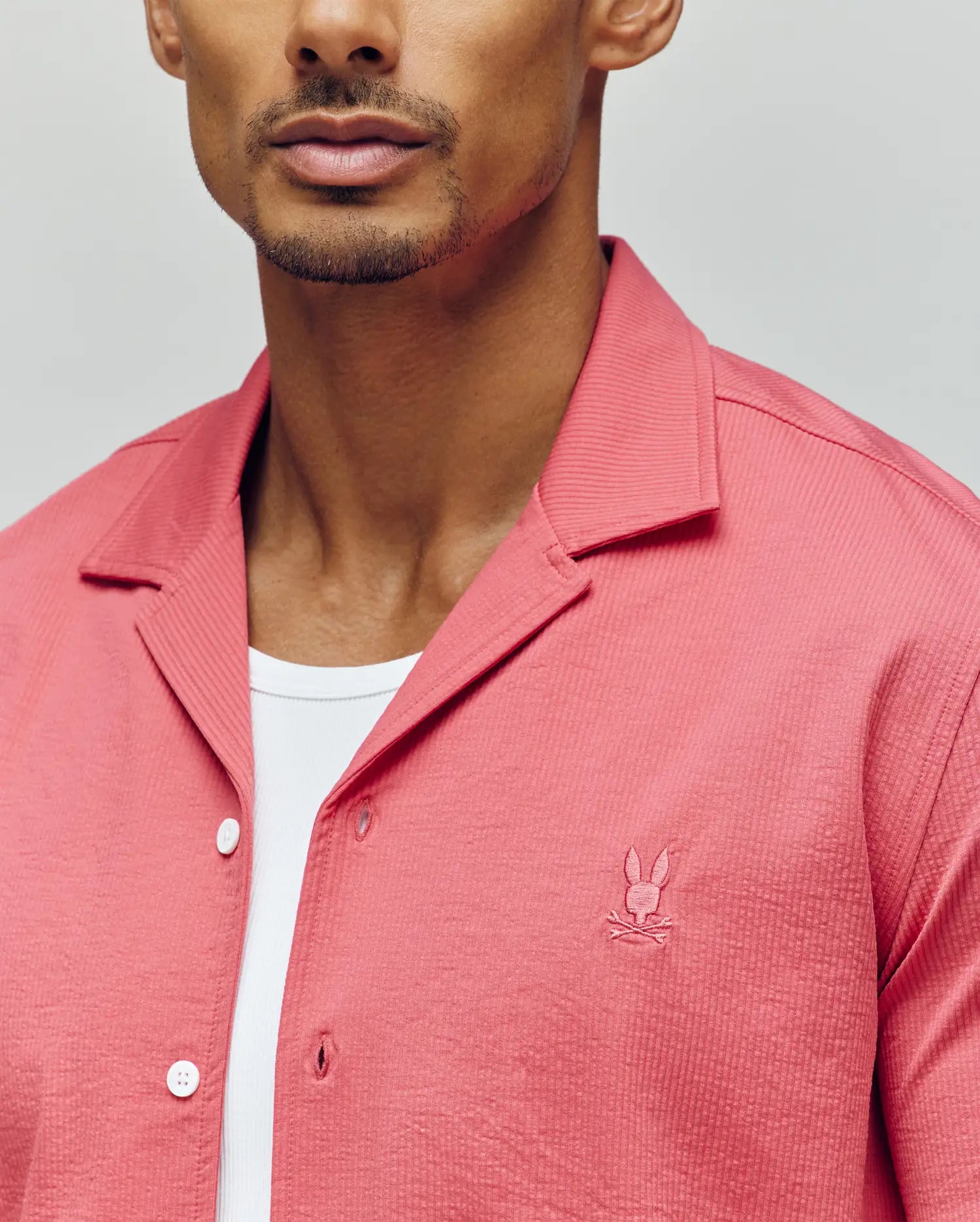 A man wears the PsychoBunny MENS JAYME KNIT SEERSUCKER CABANA COLLAR SHIRT (MT0300002) in coral pink, featuring a small bunny logo on the chest over a white tee, crafted from premium cotton blend, shown shoulders to mid-chest.