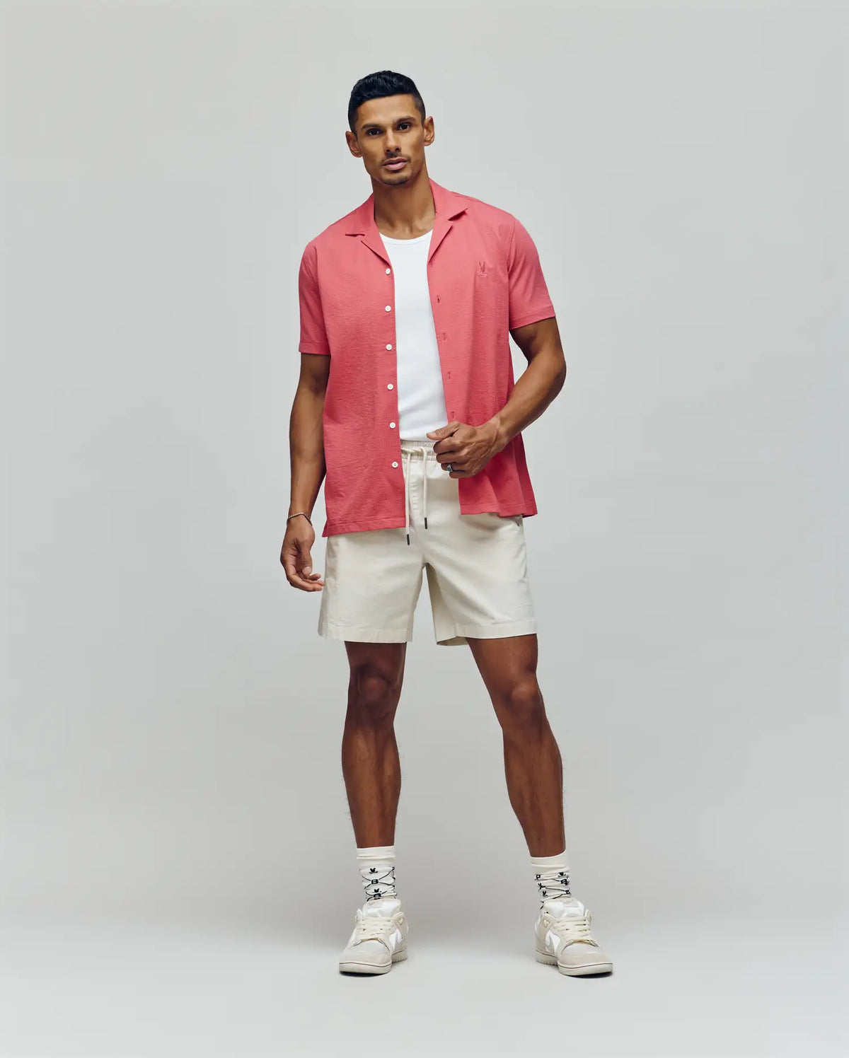 A man stands against a plain background wearing the PsychoBunny MENS JAYME KNIT SEERSUCKER CABANA COLLAR SHIRT (MT0300002) in pink over a white T-shirt, with cream drawstring shorts, patterned socks, and white sneakers.