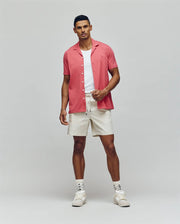 A man stands against a plain background wearing the PsychoBunny MENS JAYME KNIT SEERSUCKER CABANA COLLAR SHIRT (MT0300002) in pink over a white T-shirt, with cream drawstring shorts, patterned socks, and white sneakers.