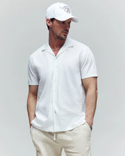 A man stands with hands in pockets, wearing the Psycho Bunny MENS JAYME KNIT SEERSUCKER CABANA COLLAR SHIRT (MT0300002), white pants, and a white baseball cap, looking to the side against a plain light gray background.