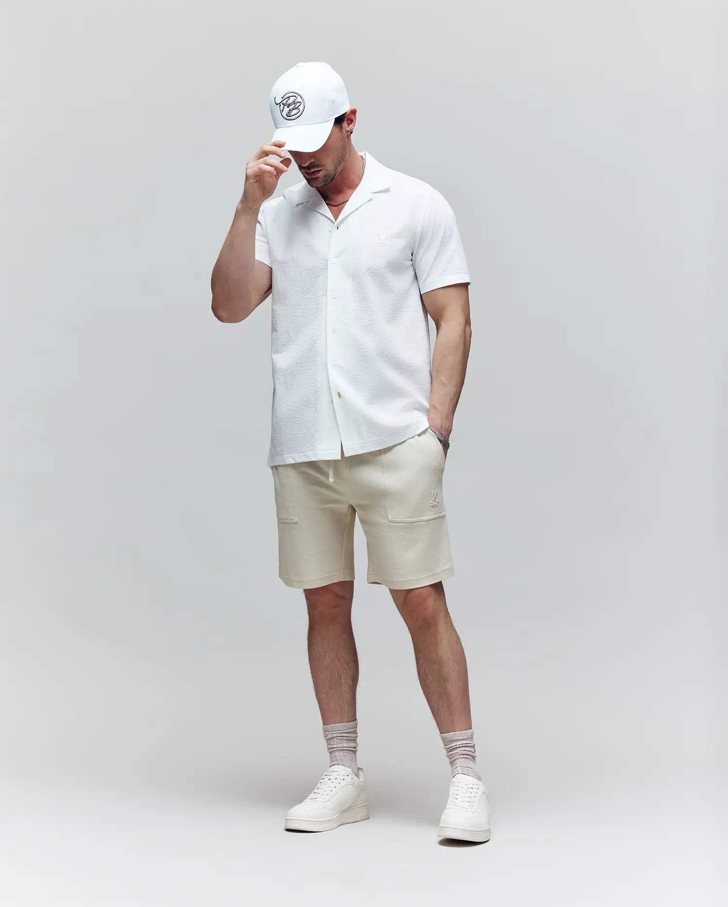 A man stands against a plain background wearing the Psycho Bunny MENS JAYME KNIT SEERSUCKER CABANA COLLAR SHIRT (MT0300002), paired with beige shorts, white sneakers, socks, and a white cap. He looks down, touching the brim of his hat.