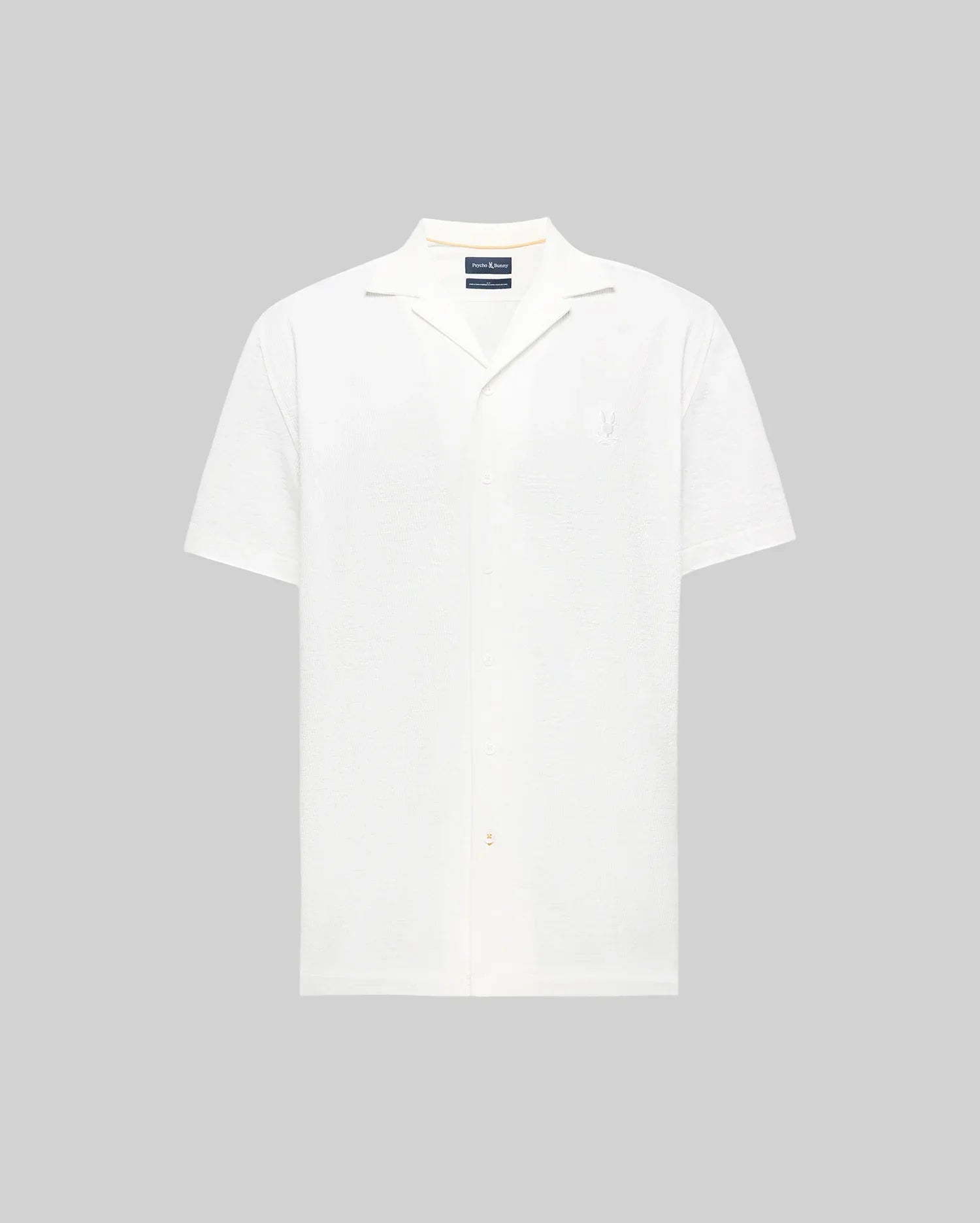 The Psycho Bunny MENS JAYME KNIT SEERSUCKER CABANA COLLAR SHIRT - MT0300002 is a white, short-sleeved, button-up shirt with a relaxed cabana collar, shown against a plain light gray background.