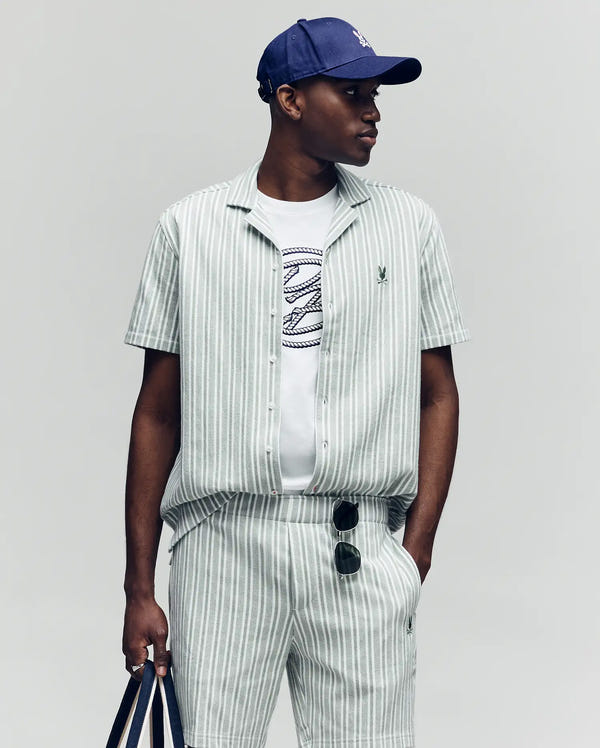 Wearing the Psycho Bunny MENS ROBERT TEXTURE STRIPE SHIRT—white and gray vertical stripes with a cabana collar—and matching shorts, they style it with a blue cap, white graphic tee, striped bag, and sunglasses hooked to their pocket.