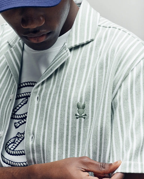 A person wears the Psycho Bunny MENS ROBERT TEXTURE STRIPE SHIRT (MT0300005), featuring light green and white stripes with an embroidered skull and bunny logo, over a white T-shirt with a blue rope design, plus a blue cap. Face is partly out of frame.