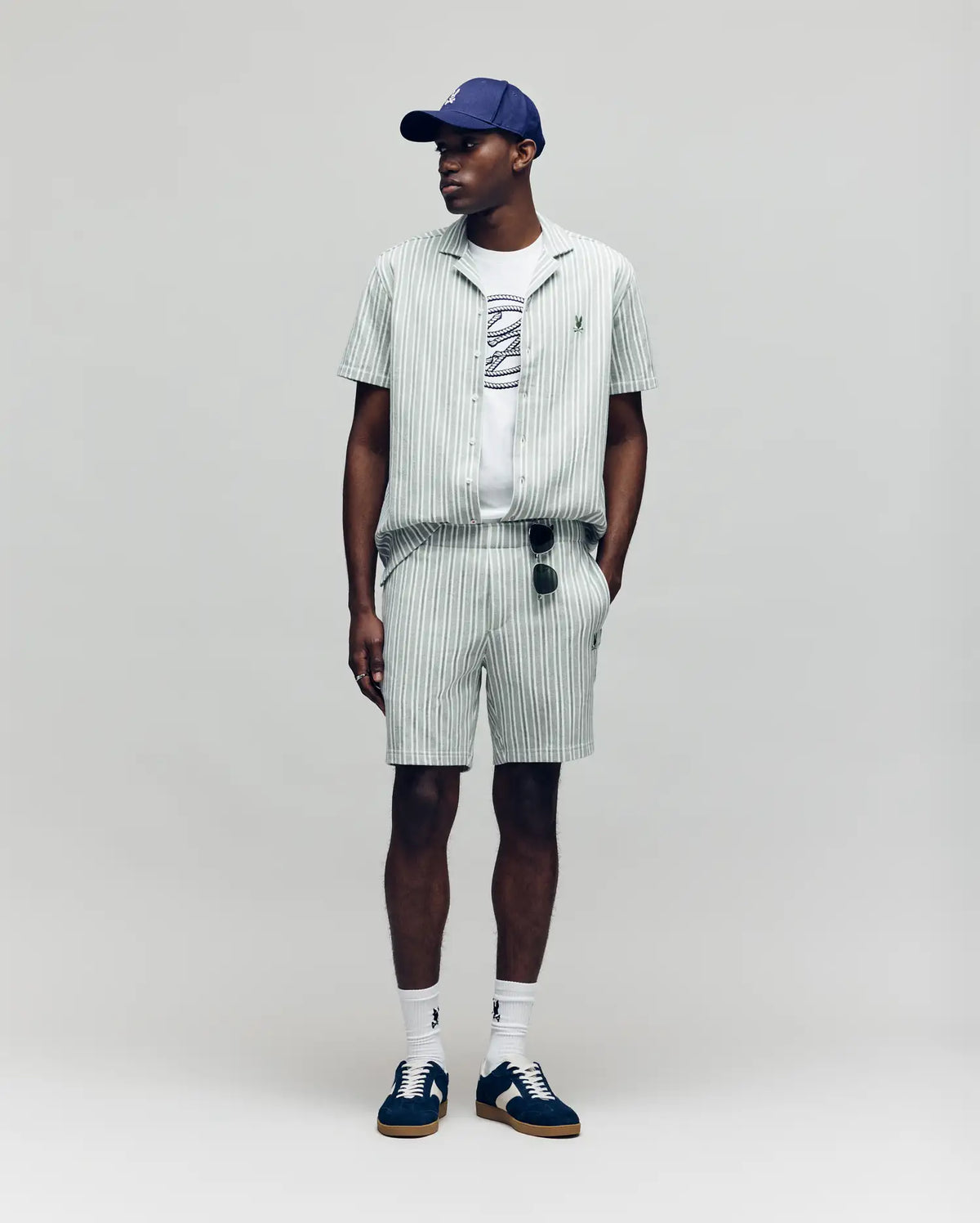 A man wears the Psycho Bunny MENS ROBERT TEXTURE STRIPE SHIRT (MT0300005) in light green with pinstripes, paired with shorts, a white graphic tee, navy cap, white socks, dark sneakers, and sunglasses hanging from his shorts. Background is plain light gray.