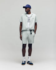 A man wears the Psycho Bunny MENS ROBERT TEXTURE STRIPE SHIRT (MT0300005) in light green with pinstripes, paired with shorts, a white graphic tee, navy cap, white socks, dark sneakers, and sunglasses hanging from his shorts. Background is plain light gray.