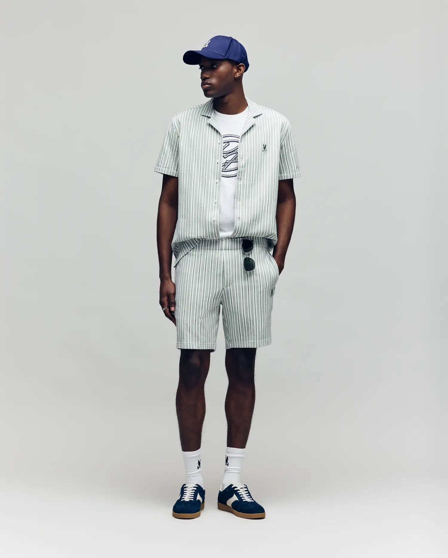 A man wears the Psycho Bunny MENS ROBERT TEXTURE STRIPE SHIRT (MT0300005) in light green with pinstripes, paired with shorts, a white graphic tee, navy cap, white socks, dark sneakers, and sunglasses hanging from his shorts. Background is plain light gray.