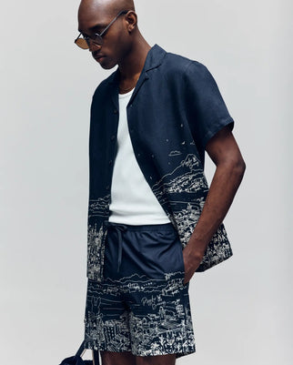 A man in Psycho Bunny’s NAXOS LINEN BLEND SHIRT - MT0300014, a dark short-sleeve shirt with white cityscape patterns, matching shorts, and sunglasses stands against a plain light background.
