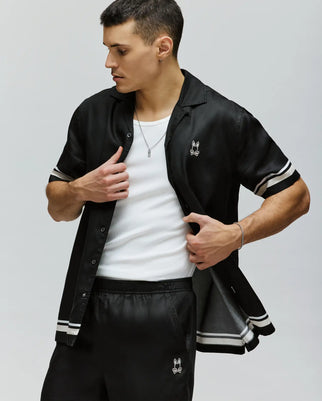 A man wears the Psycho Bunny MENS ADEN BOARDER PRINT SHIRT - MT0300015, a sustainable black short-sleeve button-up with white stripes and a small bunny logo, layered over a white tank top and paired with matching black shorts. He adjusts his shirt while looking to the side.