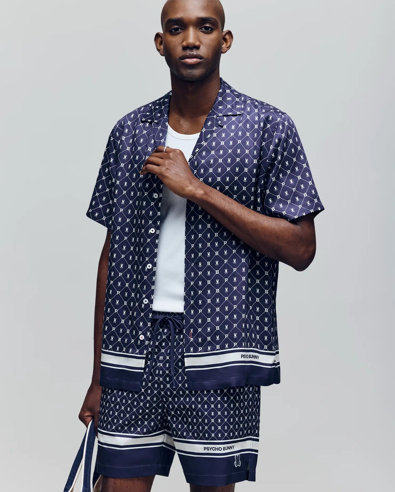 A man wears the Psycho Bunny MENS HUNTER TENCEL SHIRT (MT0300016) in navy with a white geometric pattern, matching shorts, and a white tank top. He holds a navy and white bag against a light gray background.
